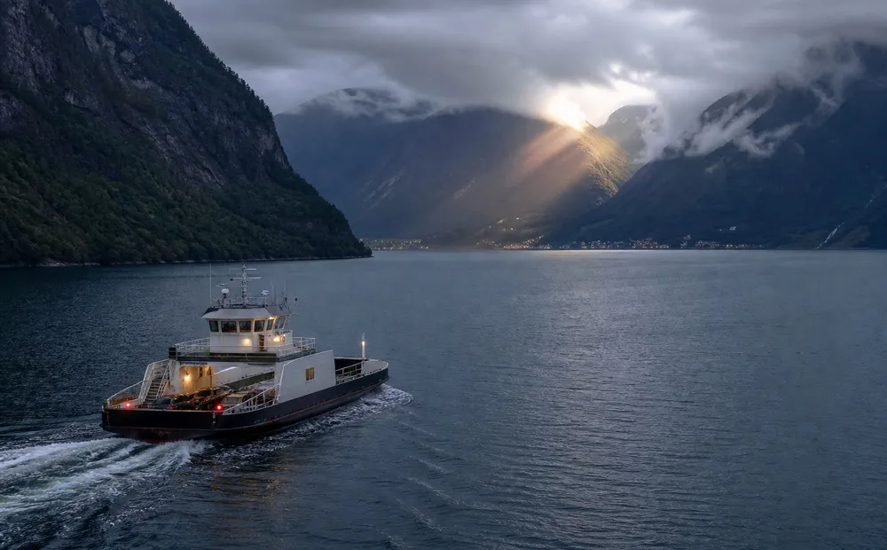 A small vehicle ferry crosses dark water between steep Norwegian fjord walls.